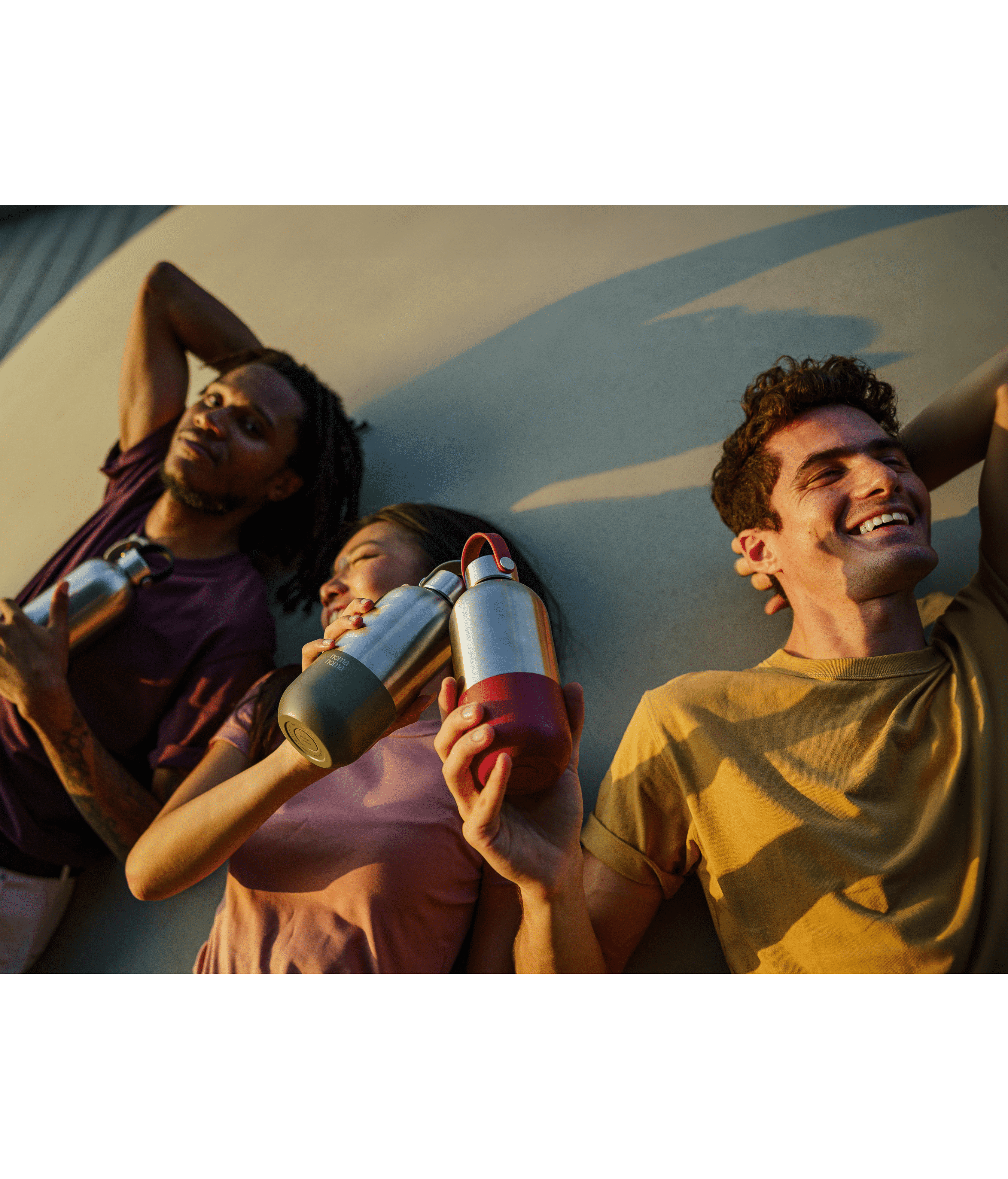 Eine Freundesgruppe lächelt und lacht in der Sonne liegend und stoßt mit den noma noma Trinkflaschen an.