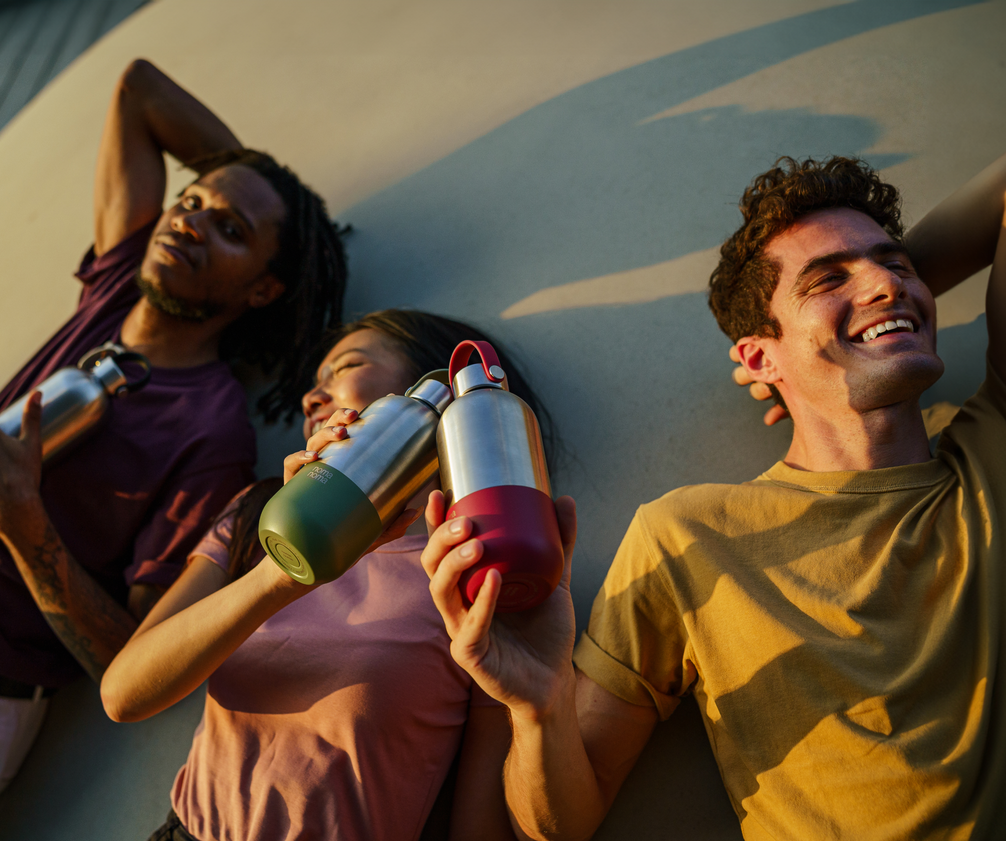 Eine Freundesgruppe lächelt und lacht in der Sonne liegend und stoßt mit den noma noma Trinkflaschen an.